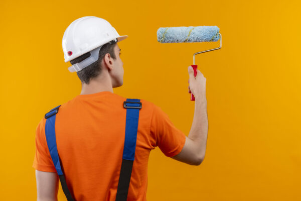young builder man wearing construction uniform and safety helmet paints the wall with a roller brush over orange isolated background
